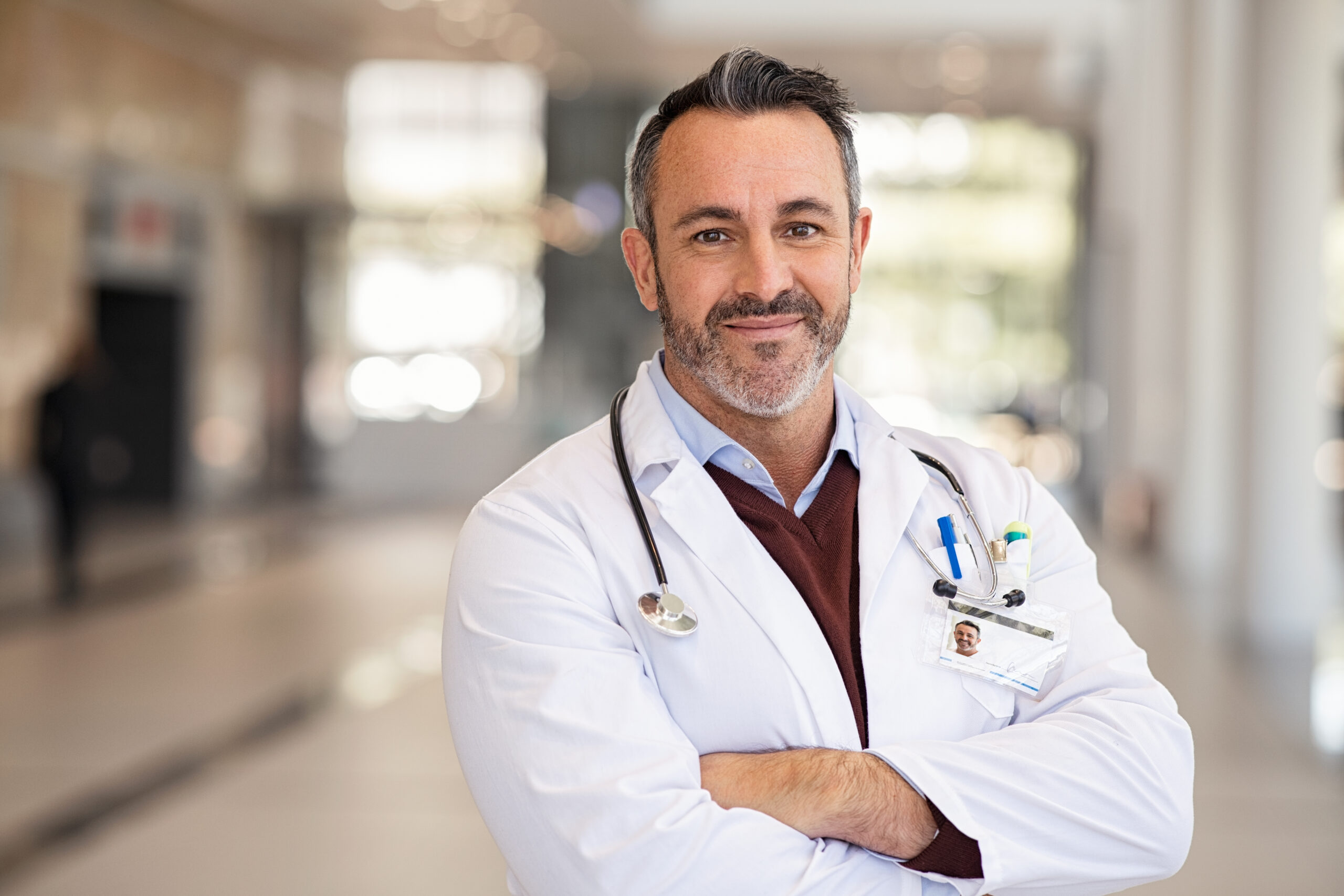 Portrait of happy doctor in labcoat and stethoscope standing with folded hands in modern hospital corridor. Successful general practitioner looking at camera with satisfaction at private clinic. Confident proud head physician standing in hospital hallway smiling with copy space.